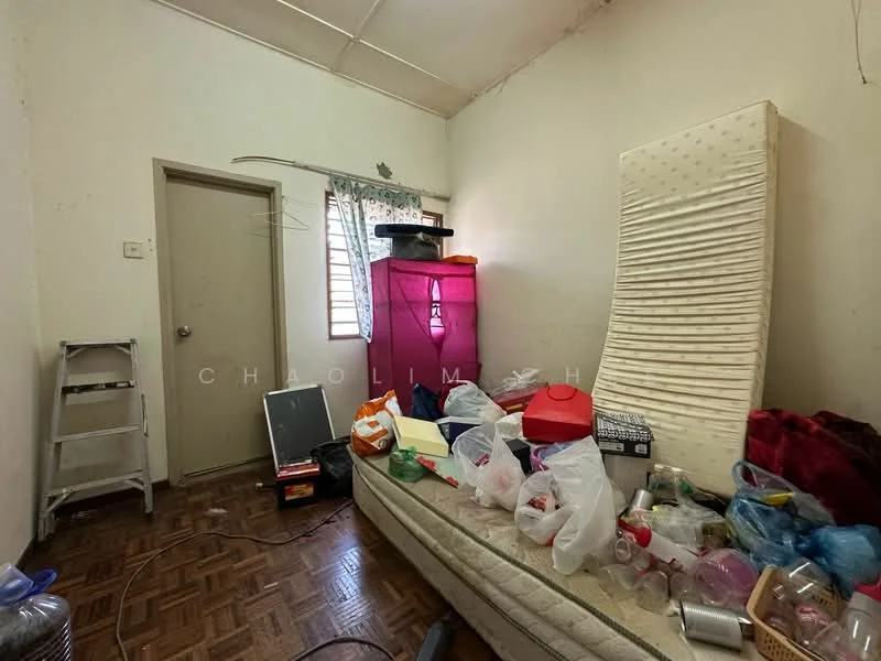 Room features: wooden flooring, white painted walls, single window with curtain, mattress on bed frame, various plastic containers, clothes in bags, folding chair, shelf unit, pink storage cabinet, miscellaneous items scattered.