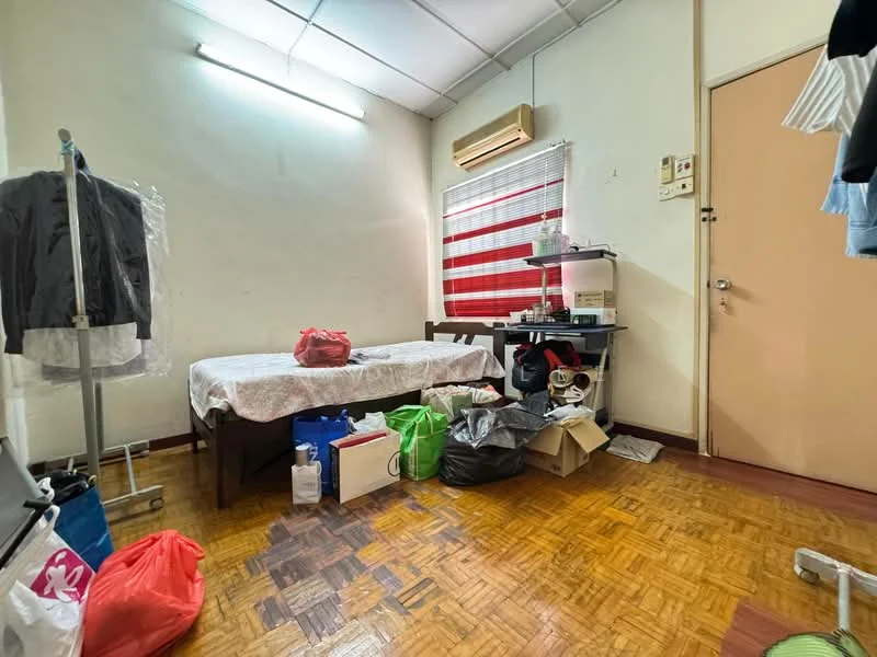 Room features: single bed with floral bedding, wooden bed frame, wooden flooring, window with red and white striped curtains, wall-mounted air conditioning unit, desk with various items on it, clothing rack with garments, plastic storage bags, cardboard boxes, garbage bags, light fixture on ceiling.