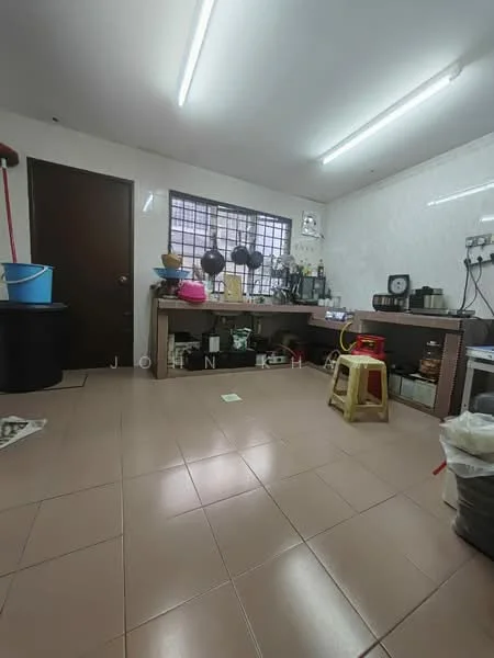 Room features: tile flooring, white painted walls, fluorescent ceiling lights, window with metal grilles, wooden door, countertop with storage underneath, various kitchen utensils and appliances, plastic stool, cleaning supplies, sink, broom, bucket; style: functional.