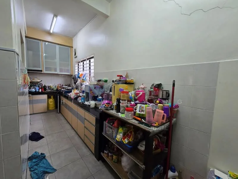 Kitchen features: L-shaped kitchen layout, grey tiled flooring, light beige cabinetry, stainless-steel sink, built-in countertop, multiple kitchen appliances visible, window with metal grille, overhead fluorescent lighting, cluttered with various kitchen items.