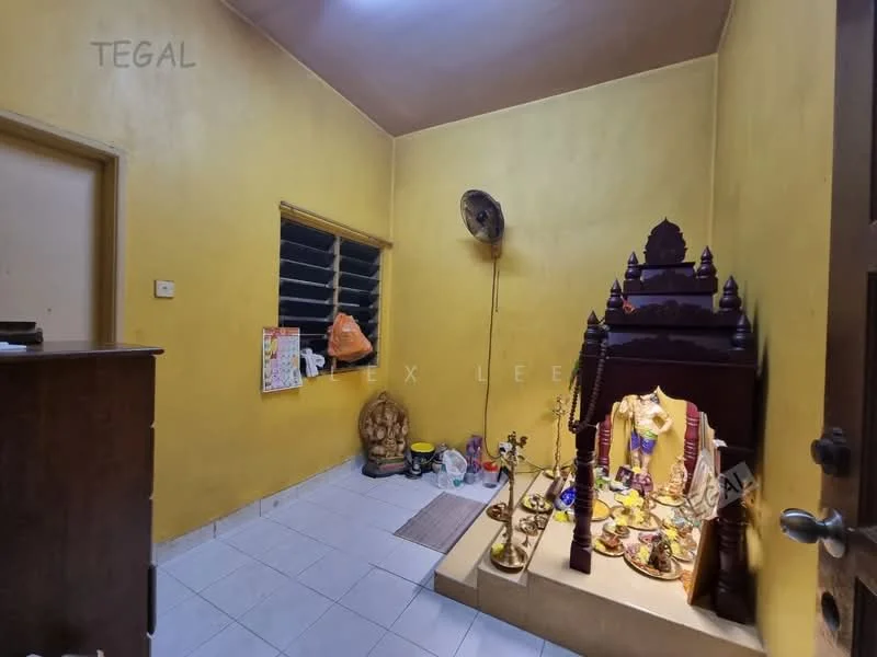 Room features: yellow painted walls, white tiled flooring, window with metal bars, wall-mounted ceiling fan, wooden door, decorative altar with offerings, small decorative items on the floor, mat at entrance.