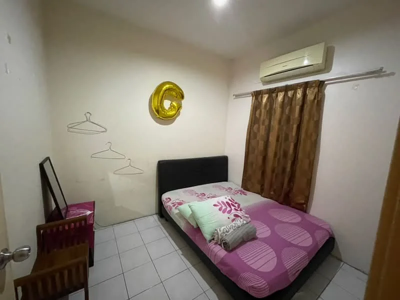 Bedroom features: queen-size bed with black frame, pink and white patterned bedding, two wooden chairs, mirror on a small table, hanging clothes rack, brown curtains, wall-mounted air conditioning unit, white painted walls, tile flooring, decorative gold balloon 'G'.