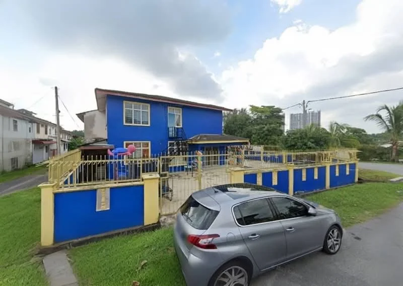 Exterior type: detached bungalow; features: blue painted facade, two-storey building, balcony with railing, manicured lawn, paved walkway, surrounded by greenery, car porch, multiple windows, stairs leading to balcony, fenced perimeter; style: modern.