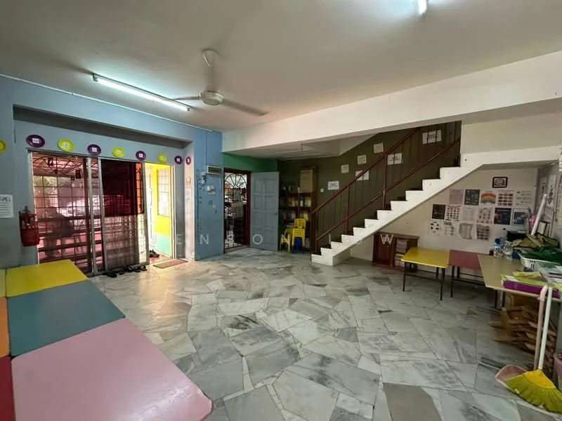 Room features: marble flooring, white walls, ceiling fan, staircase with red railing, large windows with metal grilles, open layout, multiple seating areas, colorful wall decorations, yellow chairs, tables with various materials, fire extinguisher mounted on wall.
