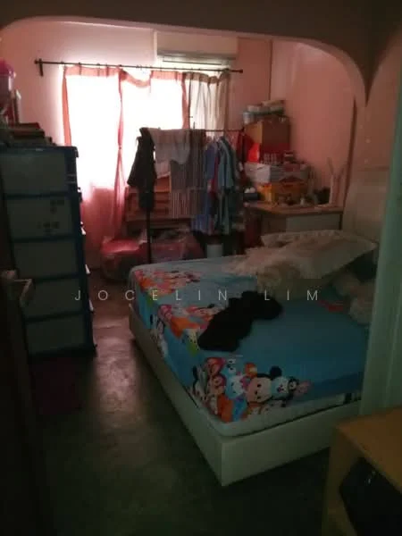 Bedroom features: queen-size bed with colorful bedding, wooden wardrobe, plastic storage drawers, window with sheer curtains, pink painted walls, concrete flooring, clothes hanging on hooks, decorative items on shelves.