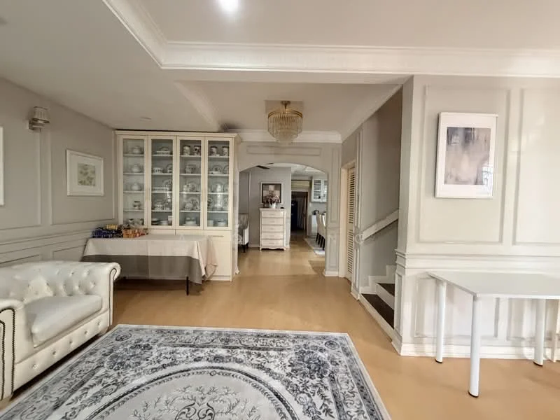 Living features: cream leather chesterfield sofa, area rug with floral pattern, wooden flooring, white painted walls with crown moulding, chandelier with crystals, glass display cabinet, decorative wall art, arched doorway leading to hallway; style: traditional. Room features: staircase with wooden steps, white painted walls, light-colored flooring.