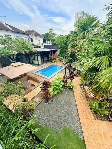 Exterior type: detached bungalow; features: infinity pool, wooden deck surrounding pool, landscaped garden with tropical plants, manicured lawn, stone-paved walkway, outdoor seating area with umbrellas, covered patio, surrounded by greenery; style: tropical-modern.