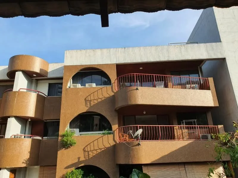 Exterior type: low-rise apartment block; features: brown painted facade, curved balconies, glass windows, metal railing on balconies, outdoor seating on balcony, satellite dish visible, greenery around the base, two-storey building, concrete structure; style: modern.