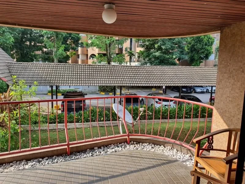 Balcony features: red metal railing, grey tiled flooring, wooden chair, decorative stone pebbles, overhead light fixture, view of landscaped garden, view of parking area, surrounding greenery; view: garden.