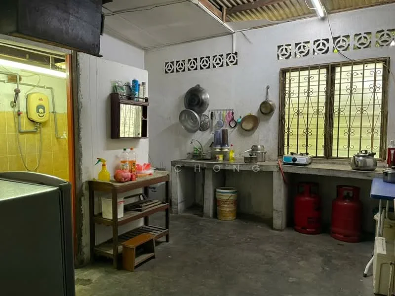 Kitchen features: concrete flooring, white painted walls, metal sink, wall-mounted gas water heater, wooden shelving unit, stainless-steel pots and pans, two gas cylinders, window with metal grilles, countertop with storage underneath, refrigerator, cleaning supplies on countertop, plastic containers, kitchen utensils, wall-mounted hooks for pots; style: functional.