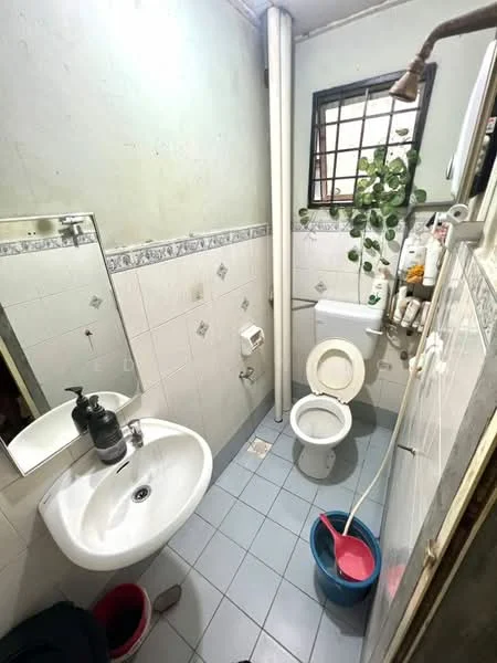 Bathroom features: white tiled walls, light blue tiled flooring, single-sink vanity with mirror, toilet, shower with handheld showerhead, plant decoration, soap dispenser, toilet paper holder, bucket, plumbing pipes visible.