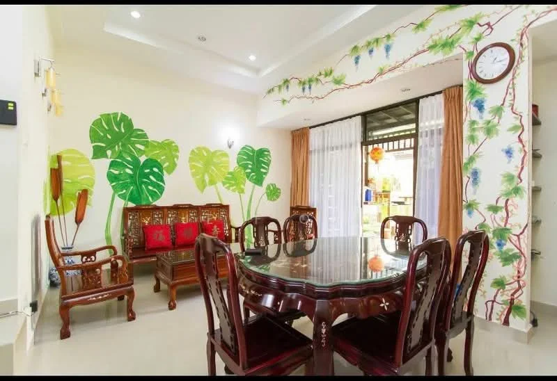 Dining features: round glass dining table, 8 wooden dining chairs with intricate carvings, decorative wall mural with green leaves, cream painted walls, recessed ceiling lights, curtains in light brown, wooden sideboard, wall clock; style: traditional; view: garden.