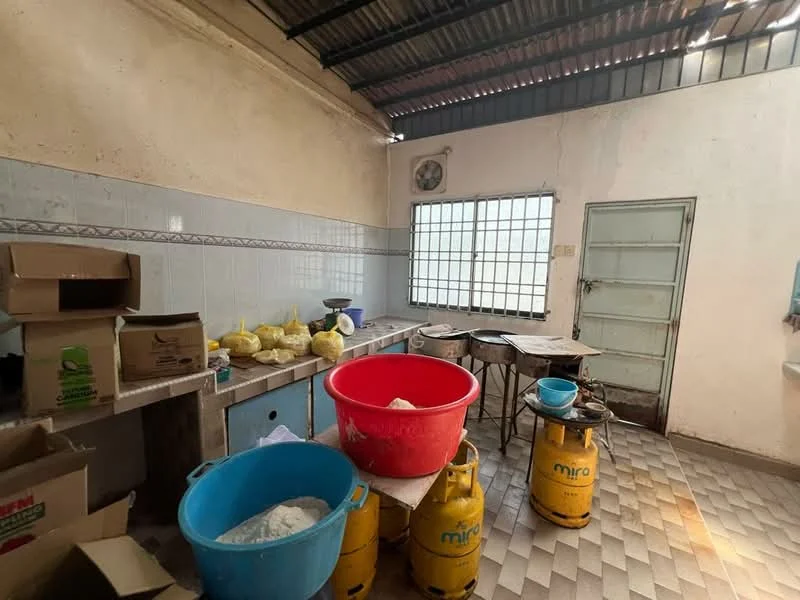 Kitchen features: blue plastic basin, red plastic basin, yellow gas cylinder, grey gas cylinder, wooden countertop, white tiled walls, grey tiled flooring, metal table, window with metal grille, boxes stacked on countertop, scale on countertop.