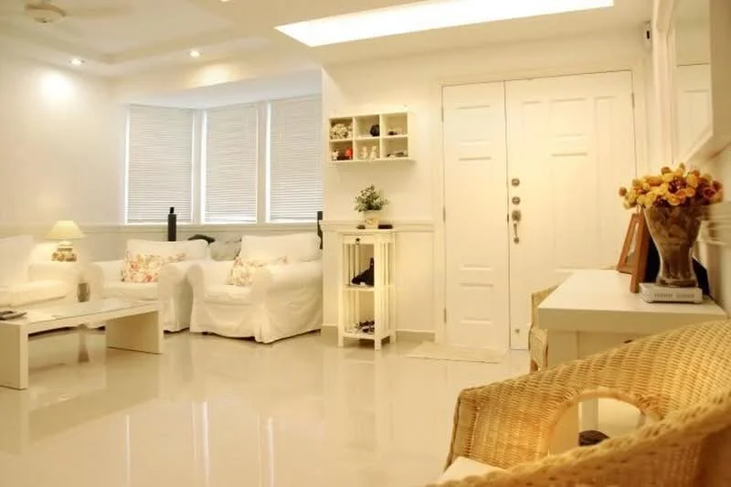 Living features: white painted walls, glossy tile flooring, tray ceiling with recessed lighting, 3-seater white fabric sofa, 2-seater white fabric sofa, coffee table, decorative wall shelf, floor lamp, window blinds, entrance door with double locks, decorative vase with flowers, wicker chair; style: modern.