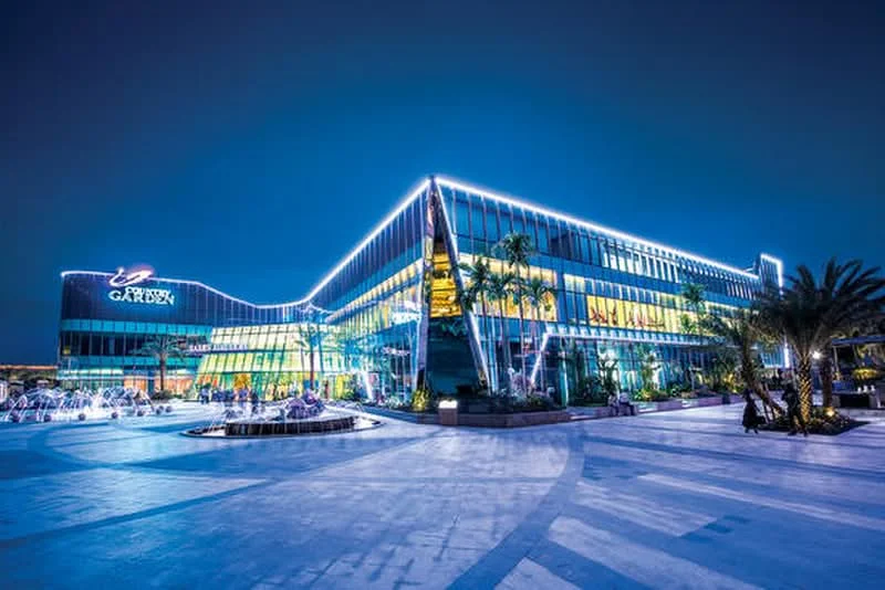 Exterior type: shopping mall; features: glass curtain wall, illuminated facade, palm trees, outdoor seating area, manicured landscaping, water feature, modern architectural design, multi-storey structure; style: modern.