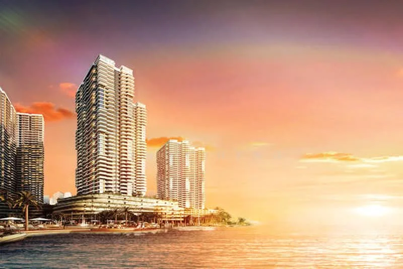Exterior type: high-rise condominium; features: glass curtain wall, multiple towers, waterfront view, sunset backdrop, surrounded by palm trees, outdoor seating area, landscaped gardens, commercial space at ground level; style: modern.