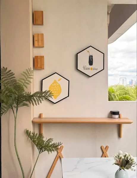 Room features: hexagonal wall art with black frames, light wood shelf, cream painted walls, decorative plant, white marble tabletop, wooden accents, natural light from window, greenery visible outside; style: modern; view: garden.