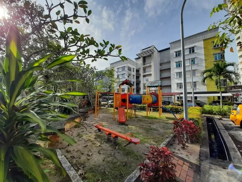 Exterior type: low-rise apartment block; features: children's playground with colorful play equipment, seesaw, surrounding greenery with shrubs, paved walkway, parking area visible, residential building facade with yellow and white colors, mature trees, sunlight filtering through leaves; style: modern.