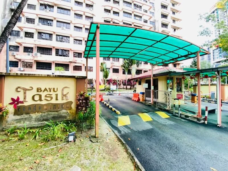 Exterior type: low-rise apartment block; features: covered car porch with green canopy, security guard house at entrance, paved driveway, landscaped garden with plants, signage with 'Bayu Tasik' name, traffic barriers, surrounding mature trees, multiple-storey building in background; style: modern.