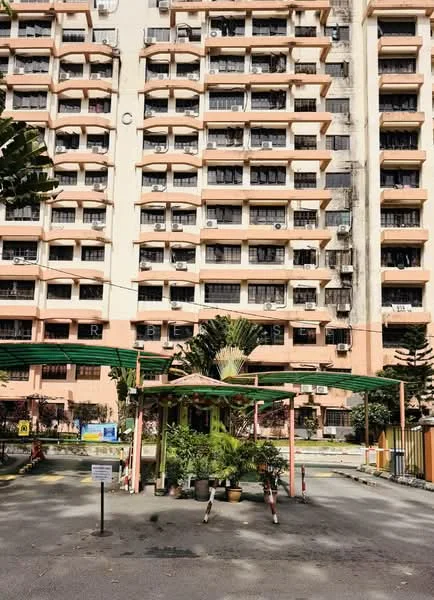 Exterior type: low-rise apartment block; features: pink painted facade, multiple floors with balconies, air conditioning units on windows, covered entrance with green awning, potted plants at entrance, manicured landscaping, parking area visible, security sign at entrance; style: modern.