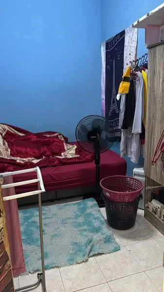 Bedroom features: blue painted walls, queen-size bed with red bedding, floor fan, textile hanging on wall, clothes rack with hanging clothes, plastic laundry basket, area rug with teal pattern, tile flooring, built-in wardrobe.