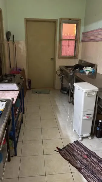Kitchen features: white tiled walls, cream tiled flooring, stainless-steel countertop, stainless-steel sink, gas stove with 4 burners, metal shelving unit, white refrigerator, window with wooden frame, door leading to another area, textile rug on floor.