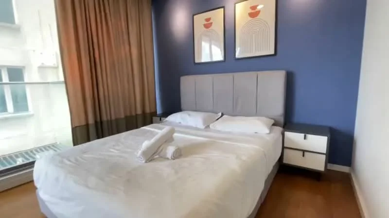 Bedroom features: king-size bed, upholstered headboard, white bedding, 2 white towels, nightstand with 2 drawers, framed artwork on walls, floor-to-ceiling windows, curtains, wooden flooring, blue accent wall; style: modern.