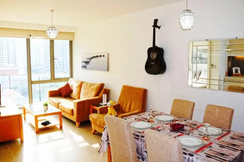 Living features: brown fabric sofa, 2 beige armchairs, wooden coffee table, dining table with 6 chairs, decorative tablecloth, guitar mounted on wall, framed mirror, pendant lighting, floor-to-ceiling windows, city view, light-colored tile flooring, wall art, decorative vase; style: modern; view: city.