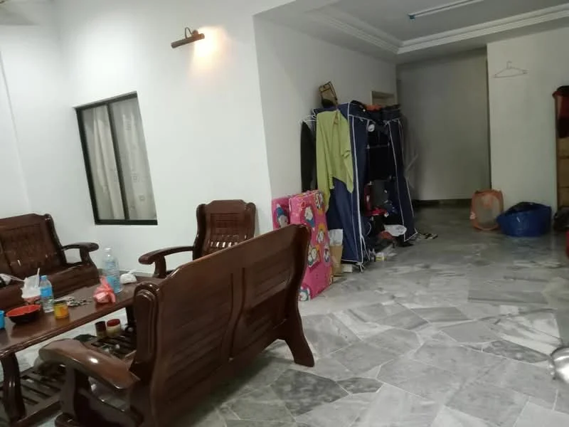 Living features: grey marble flooring, white painted walls, wooden furniture, wooden coffee table, two wooden chairs, clothes drying rack, various bags and items in the corner, window with sheer curtains, ceiling with recessed lighting.