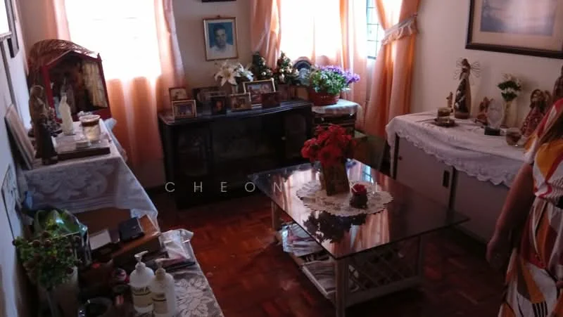 Living features: wooden flooring, cream painted walls, large windows with sheer curtains, glass coffee table, wooden side table, decorative tablecloths, framed photographs on display, potted plants, various decorative figurines, vase with flowers, wall-mounted artwork, shelves with decorative items; style: traditional.