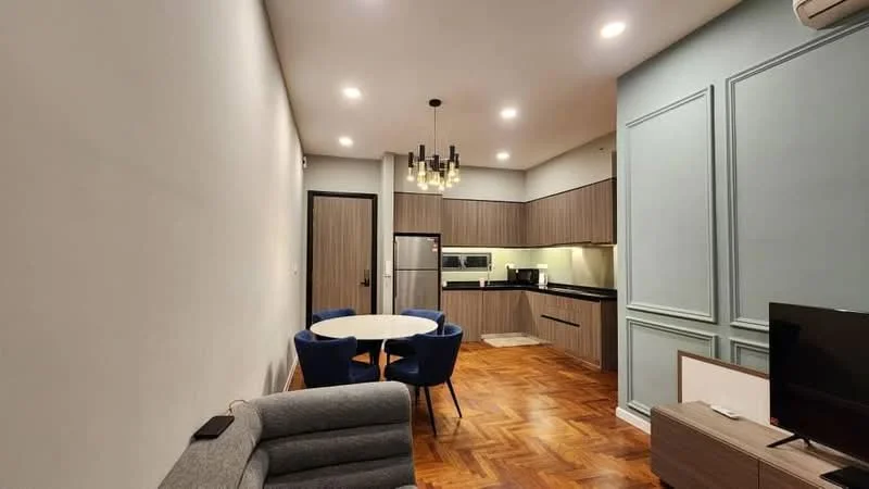 Living features: grey fabric sofa, round dining table, 4 blue upholstered dining chairs, built-in kitchen cabinetry in wood finish, black countertop, stainless-steel fridge, built-in oven, ceramic cooktop, pendant lighting, recessed ceiling lights, parquet wood flooring, white painted walls, wall-mounted TV; style: modern.