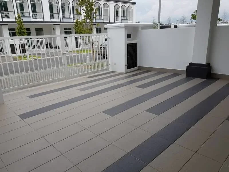 Exterior type: linked terrace house; features: paved flooring with alternating grey and beige tiles, white perimeter wall, fenced area with metal railing, outdoor wall-mounted ventilation unit, landscaped area with small trees, open space in front of the house; style: modern.