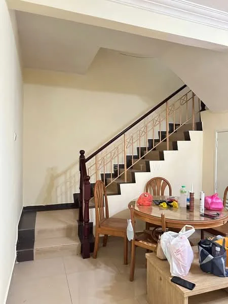 Dining features: round wooden dining table, four wooden dining chairs, cream painted walls, tile flooring, staircase with wooden railing, mirror on wall, decorative items on table, plastic bags on floor. Room features: staircase with black steps, light-colored wall, ceiling with crown moulding.