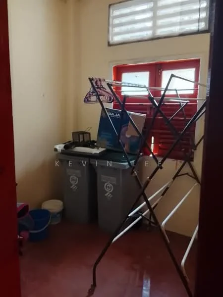 Laundry features: yellow painted walls, red window frames, window with horizontal bars, clothes drying rack, two grey trash bins, plastic blue bucket, small table, flooring with red tiles.