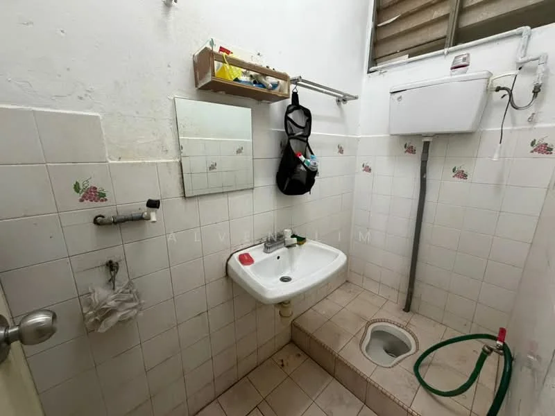 Bathroom features: white tiled walls, cream tiled flooring, sink with wall-mounted faucet, mirror above sink, toilet bowl, hose on the floor, shelf with toiletries, window for natural light.
