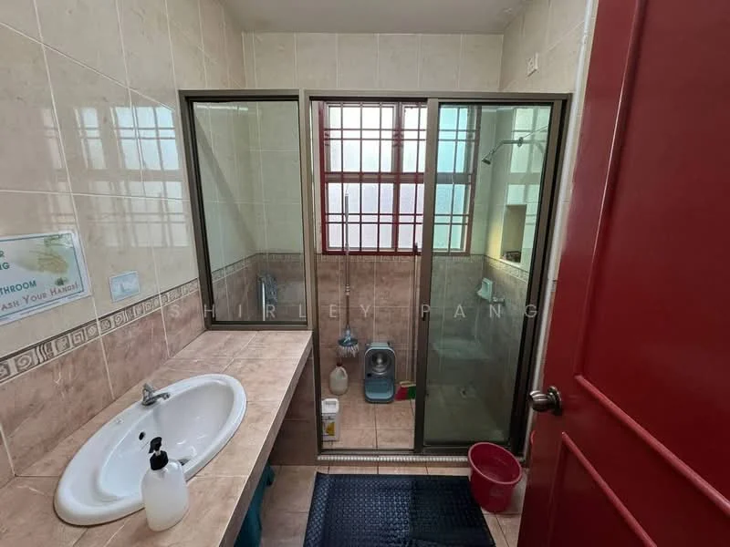 Bathroom features: single-sink vanity with white cabinet, chrome basin mixer faucet, mirror above sink, glass shower enclosure, toilet, tile flooring, cream tiled walls, window for natural light, red door, plastic bucket, cleaning supplies, floor mat.