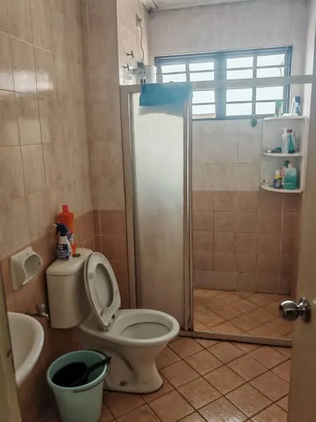 Bathroom features: light beige tiled walls, light beige tiled flooring, shower cubicle with glass door, toilet, sink with wall-mounted faucet, window with metal grilles, shower shelf with toiletries, cleaning supplies in bucket.
