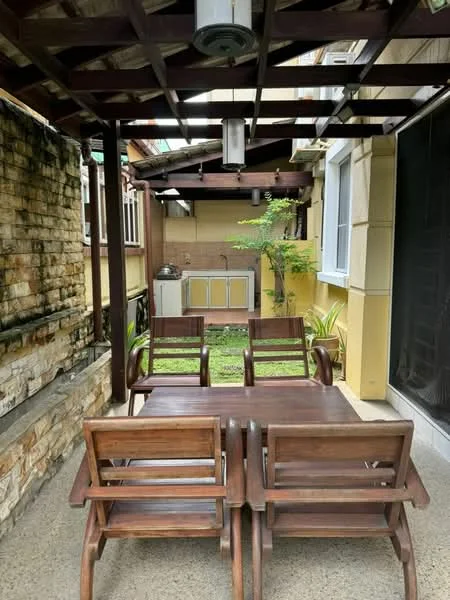 Room features: wooden dining table, four wooden chairs, exposed wooden beams, stone wall, green grass area, outdoor kitchen with cabinetry, wall-mounted light fixture, paved flooring; style: modern; view: garden.