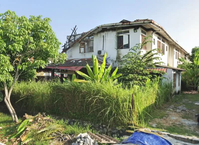 Exterior type: semi-detached house; features: two-storey structure, red tiled roof, white painted facade, exposed brick wall, overgrown garden, mature trees, tall grass, concrete pathway, air conditioning unit visible, wooden window frames, balcony on upper floor; style: traditional.