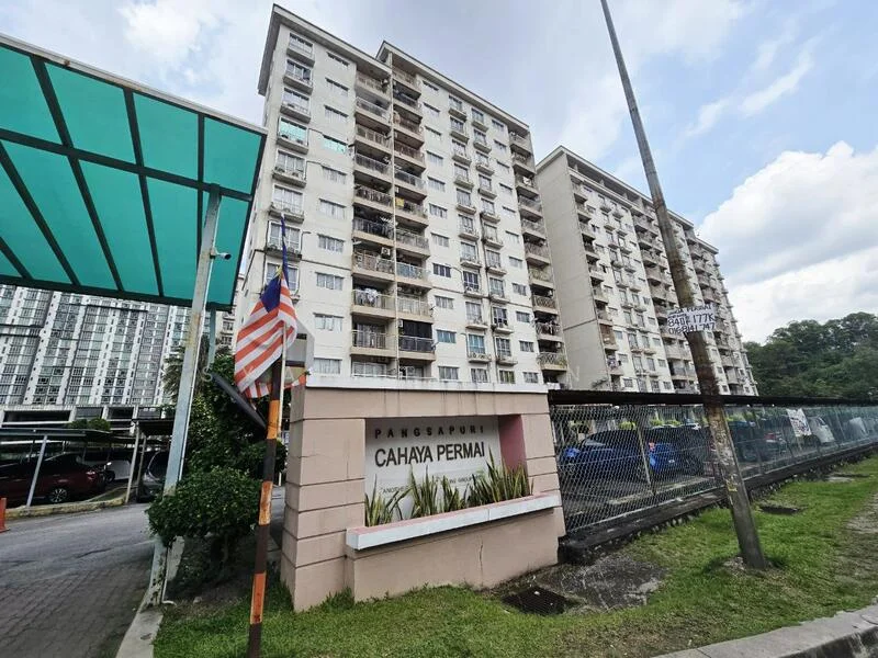 Exterior type: mid-rise condominium; features: cream painted facade, balconies visible on upper floors, landscaped garden area, signage with 'Cahaya Permai', surrounding greenery, parking area visible, fenced perimeter, flagpole with Malaysian flag; style: modern.