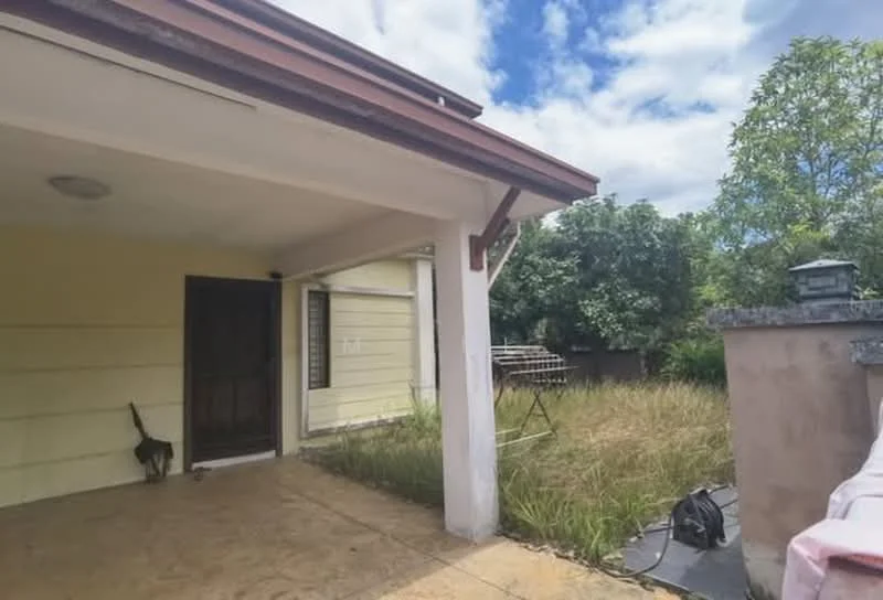 Exterior features: single-storey structure, yellow painted facade, wooden door, concrete flooring, overhang roof, surrounding greenery, unmaintained garden area, visible outdoor equipment, partially cloudy sky.