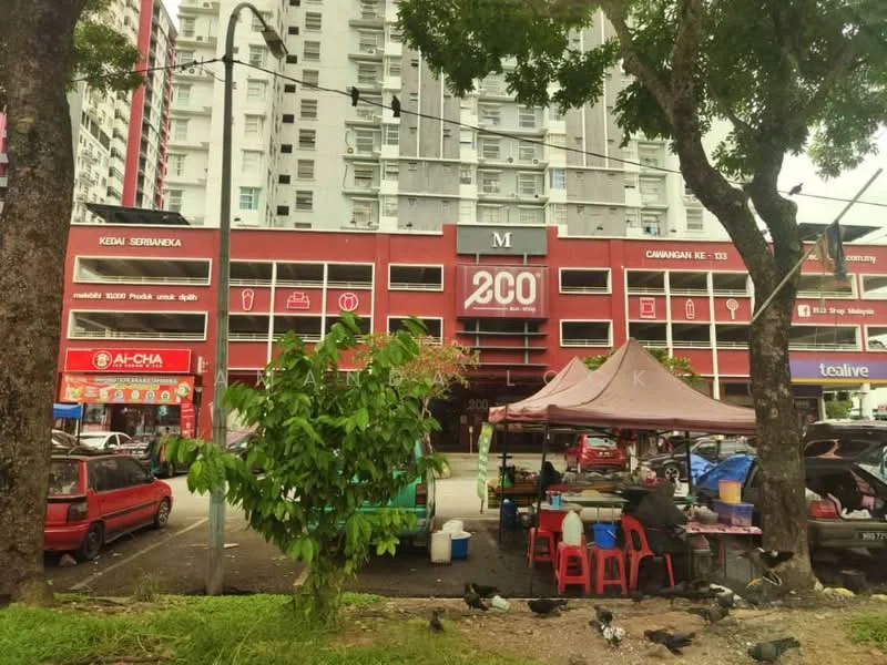 Exterior type: mid-rise condominium; features: red painted facade, ground floor retail spaces, signage for various shops, parking area in front, trees providing shade, tent structure for vendors, multiple vehicles parked, urban setting, visible power lines; style: modern.