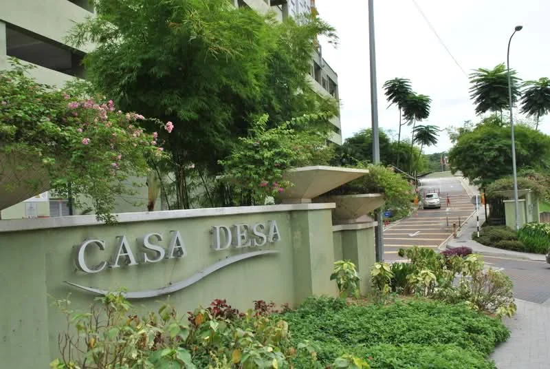 Exterior type: low-rise apartment block; features: green painted facade, landscaped garden with flowering plants, paved walkway, signage with 'CASA DESA', surrounding greenery with trees, sloped driveway, parking area visible, street lighting; style: modern.