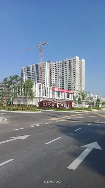 Exterior type: high-rise condominium; features: glass curtain wall, white painted facade, recessed balconies, signage at entrance, surrounded by landscaped gardens, construction crane visible, multi-storey structure, manicured lawn, paved walkway, urban context with road access; style: modern.
