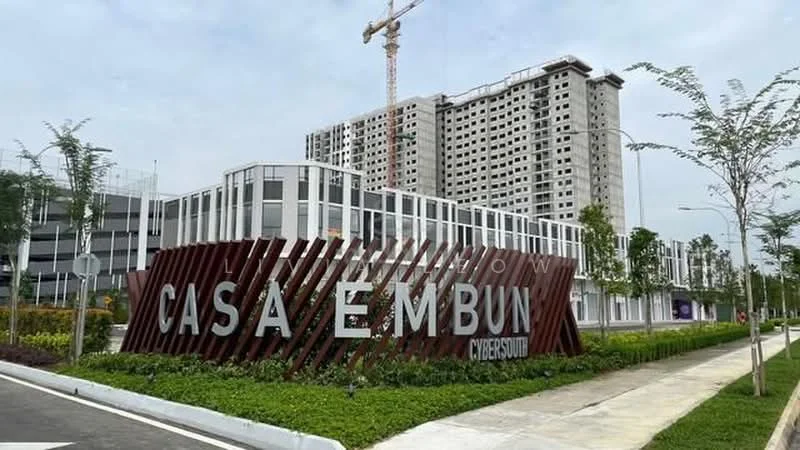 Exterior type: mid-rise condominium; features: modern facade with glass and concrete, landscaped garden with shrubs, paved walkway, signage with 'CASA EMBUN', construction crane in background, surrounding greenery, multiple storeys visible, parking area, urban setting; style: modern.