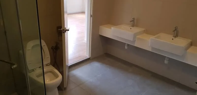 Bathroom features: two wall-mounted sinks, chrome basin mixers, one-piece toilet, glass shower enclosure, grey tile flooring, light beige wall tiles, natural light from door, minimalist design; style: modern.
