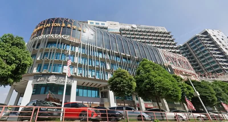 Exterior type: mixed-use development; features: glass curtain wall facade, metal cladding, projecting balconies, signage on facade, multiple storeys, landscaped greenery in front, parking area with cars, surrounded by mature trees; style: modern.