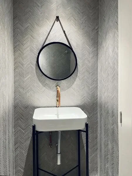 Bathroom features: white ceramic sink, gold faucet, black metal frame, chevron patterned wall tiles in grey, round mirror with black frame, wall-mounted plumbing, minimalist design, no visible storage, neutral color palette; style: modern.