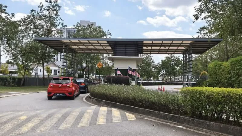 Exterior type: main entrance gate; features: modern gate structure, covered entrance with roof, security booth, landscaped greenery, manicured hedges, paved driveway, traffic signage, multiple flags displayed, surrounding trees, open-air design; style: modern.