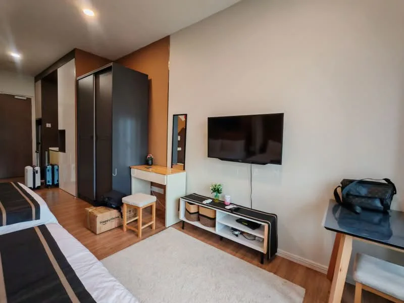 Room features: wooden flooring, painted walls in light beige, black built-in wardrobe, wall-mounted flat-screen TV, white TV console with storage, small desk with chair, decorative plant on desk, light-colored area rug, suitcases on the floor, mirror on the wall, natural light from window; style: modern.